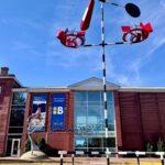 A tall colorful metal kinetic sculpture sits on the lawn of the Biggs Museum. The Museum façade and a sculpture of a twee with birds swirling into the atrium is seen in the background.